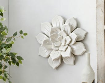 May include: A white ceramic wall sculpture shaped like a blooming succulent or flower with layered, rounded petals. The piece features a smooth, glossy finish and is mounted centrally on a plain, light-colored wall surface.