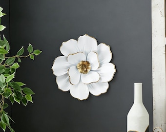 May include: White metal flower wall decor with gold trim and a gold center. The large, three-dimensional floral design is mounted on a dark gray wall. The petals have a slight curve, adding depth to the piece.