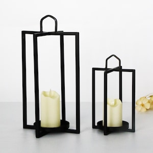 May include: Two black metal candle lanterns of different sizes. Each lantern has a square frame with a handle at the top and a circular base. A cream-colored candle sits inside each lantern, creating a warm glow.