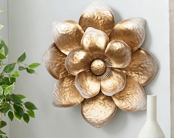 May include: A gold-colored metal wall sculpture shaped like a blooming flower with layered, hammered petals. The center features a small, raised circular disk surrounded by thin, radiating metal spokes, creating a textured floral design.