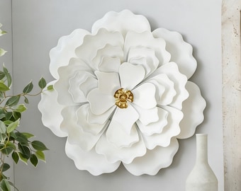 May include: A wall-mounted metal flower sculpture featuring layered, scalloped white petals arranged in a circular pattern. The center of the flower is composed of smaller, metallic gold petals that contrast with the matte white outer layers.