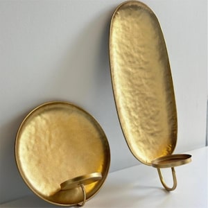 May include: Two gold-colored metal candle holders. One is round, and the other is an elongated oval shape. Both have a textured surface and a small, circular holder for a candle. The candle holders are made of metal.