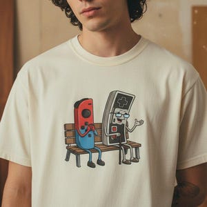 Könnte beinhalten: Cremefarbenes T-Shirt mit einer Cartoon-Illustration von zwei Videospiel-Controllern, die auf einer Bank sitzen. Ein Controller ist rot und blau, der andere grau und schwarz. Die Controller scheinen sich zu unterhalten.