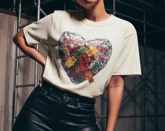 May include: Cream-colored t-shirt featuring a heart-shaped graphic filled with colorful gummy bears. The graphic is wrapped in a silver foil design. The model is wearing black leather pants.
