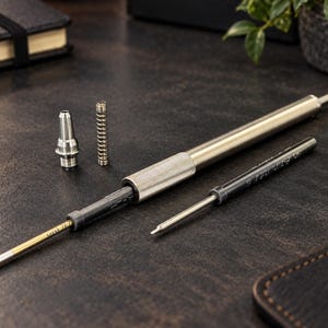 May include: A disassembled silver metal pen with a knurled grip section, a separate metal spring, a conical tip component, and two ink refills. One refill is inserted into the pen body, while the other lies alongside the main barrel.