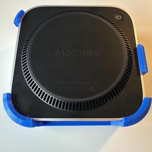 May include: A black Mac Mini computer with a silver border, held in place by a blue 3D-printed bracket. The computer has a power button and the words "Mac mini" are visible. The bracket is designed to hold the computer securely.