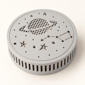 May include: A circular light grey device featuring a perforated top surface with a cutout design of a planet with rings, stars, and a constellation. The side of the cylindrical object has vertical ventilation slots around the entire perimeter.