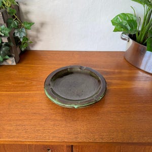May include: A round ceramic ashtray featuring a dark brown, glossy center basin and a textured, mottled green rim. The circular design includes three evenly spaced notches along the outer edge, providing a functional structure for resting items.