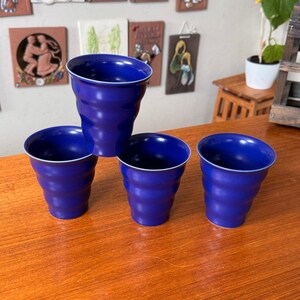 May include: A set of four cobalt blue ceramic cups with a distinct rippled, wavy texture along the exterior walls. Each cup features a wide, open circular rim and a tapered base, showcasing a consistent deep blue matte finish throughout the set.