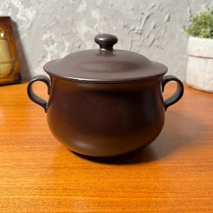 Può includere: Una pentola in ceramica marrone scuro con coperchio abbinato. La pentola ha due manici e un piccolo pomello sul coperchio. La pentola è appoggiata su una superficie di legno.