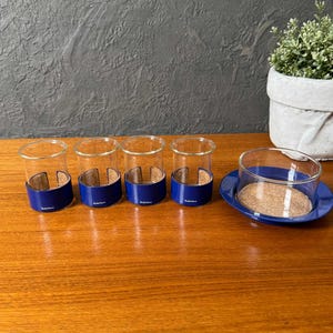 May include: A set of five clear glass tumblers with a blue base and a cork-like insert. One tumbler sits on a matching blue saucer. The glasses are arranged on a wooden surface, with a small potted plant in the background.