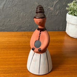 Puede incluir: Figura de cerámica de una mujer con un diseño estilizado. La figura tiene un cuerpo color melocotón, un moño marrón oscuro y una falda blanca con rayas verticales azules y grises. La figura es un objeto decorativo.