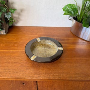 May include: A round ceramic ashtray featuring a glossy dark brown outer rim and a textured, speckled golden-brown center basin. The design includes three evenly spaced notches along the edge, each accented with a light cream and yellow glaze.