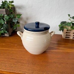 Puede incluir: Un recipiente de cerámica color crema con textura acanalada horizontal, dos asas laterales y una tapa azul oscuro. La tapa tiene un pequeño pomo central y una muesca para una cuchara. Un fino patrón de líneas onduladas rodea el borde superior.