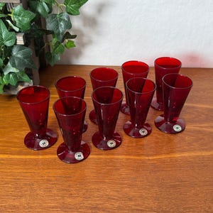 Puede incluir: Un conjunto de nueve copas de licor de cristal rojo rubí con forma cónica y base circular. Cada copa presenta una pequeña pegatina redonda en la base con la letra R impresa en negro sobre un fondo blanco, dispuestas en un grupo.