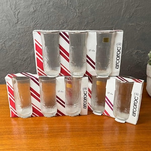 Könnte beinhalten: Ein Set aus acht transparenten, strukturierten Trinkgläsern, jeweils mit einem Fassungsvermögen von 37 cl, in der Originalverpackung. Die Verpackung hat ein rot-weißes Streifendesign und das "arcoroc"-Logo.