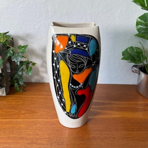 May include: A tall, off-white ceramic vase featuring a central black panel decorated with a stylised line drawing of a figure. The design includes vibrant, abstract shapes in yellow, orange, red, and blue, accented by white dotted lines and patterns.