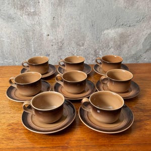 May include: A collection of eight vintage coffee cups and saucers. The cups and saucers feature a brown and tan colour scheme with a gradient effect. The cups have a rounded shape with a handle, and the saucers are circular with a dark brown rim.