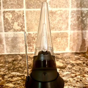 Puede incluir: Un dispositivo de vidrio negro y transparente con una parte superior cónica y una base redondeada. Una fina herramienta de metal plateado está colocada a un lado. El dispositivo está sobre una superficie marrón moteada.