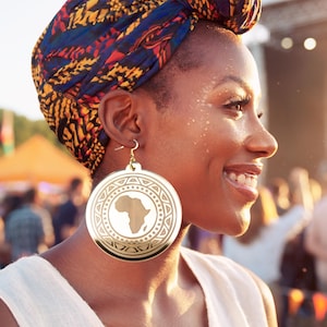 May include: A woman wearing large gold-colored earrings with an African continent design. The earrings are circular with a detailed border. She is wearing a patterned head wrap and smiling. The background is blurred, suggesting an outdoor event.