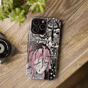 May include: A black and white phone case featuring an anime-style illustration of a character with pink hair and a distressed expression. The case has a detailed design with swords and geometric shapes. The phone case is on a wooden surface.