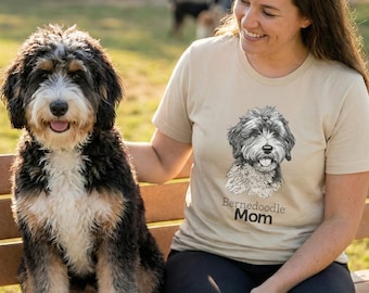 May include: A Bernedoodle dog sits on a park bench next to a person wearing a tan t-shirt. The t-shirt features a black and white illustration of a Bernedoodle and the words "Bernedoodle Mom". The dog has black, brown, and white fur.