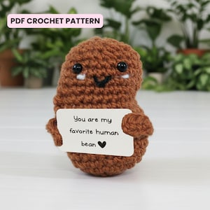 Può includere: Una figura a forma di fagiolo all'uncinetto marrone con occhi neri e una bocca sorridente. Tiene in mano un biglietto bianco con la scritta "You are my favorite human bean" con un cuore nero. Il testo "PDF CROCHET PATTERN" è in alto.