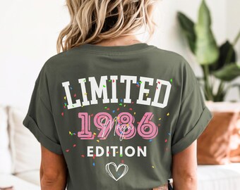 May include: An olive green t-shirt featuring the text "LIMITED 1986 EDITION" in white and pink, with confetti and a heart graphic. The shirt is worn by a person with blonde hair.