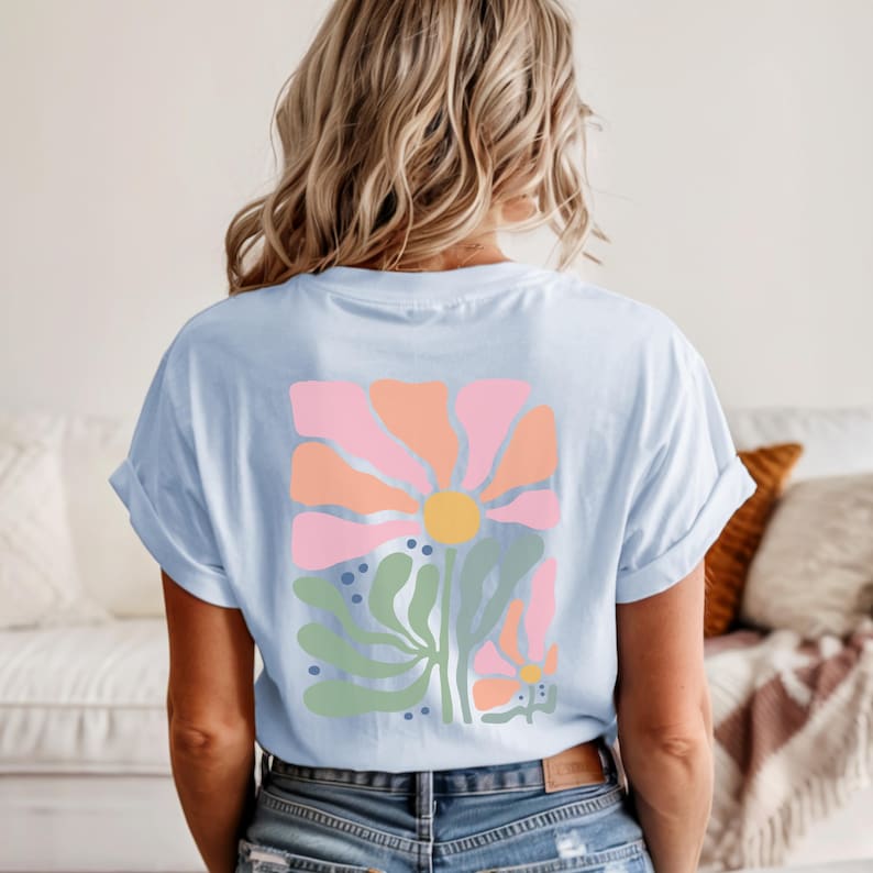 May include: A light blue t-shirt featuring a large graphic on the back. The design displays a stylised flower with peach and pink petals, a yellow centre, and sage green leaves, accented by small dark blue dots on a light blue background.
