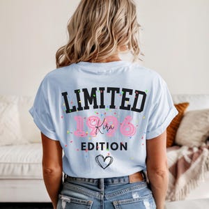 May include: Light blue t-shirt with the text "LIMITED 1986 Kira EDITION" in black and pink, with confetti and a heart graphic. The shirt is worn by a person with blonde hair.