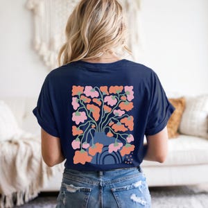 May include: Navy blue t-shirt featuring a floral design on the back. The graphic includes a vase with pink and orange flowers, green stems, and a blue arch. The shirt is made of soft material and has rolled-up sleeves.