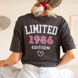 May include: Dark gray t-shirt with the text "LIMITED 1986 EDITION" in white and pink. A white heart graphic is below the text. The shirt is a casual style.