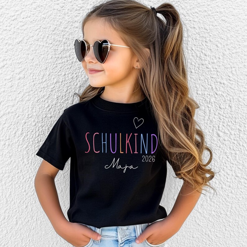 May include: A black short-sleeved t-shirt featuring the text "SCHULKIND" in multicolored letters, a small white heart outline, the name "Maja" in white script, and the year "2026" printed in white on the front of the garment.