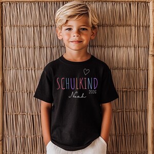 May include: A black short-sleeved t-shirt featuring the word SCHULKIND in multicolored letters, a small white heart outline, the year 2026, and the name Noah in white script. The shirt has a crew neckline and a standard fit for casual wear.