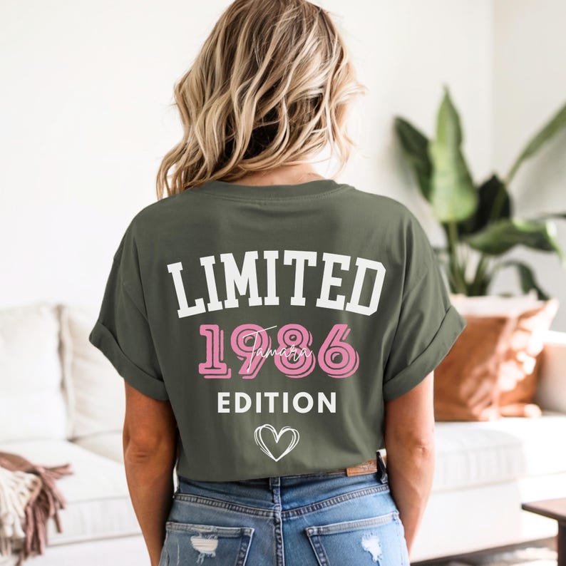May include: Olive green t-shirt with the text "LIMITED 1986 January EDITION" in white and pink. A white heart graphic is below the text. The shirt is worn by a person with blonde hair.