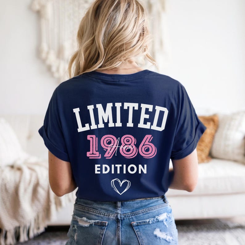 May include: Navy blue t-shirt with the text "LIMITED 1986 EDITION" in white and pink, and a small heart graphic. The shirt is worn by a person with blonde hair and blue jeans.