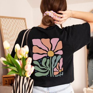May include: Black t-shirt with a large floral design on the back. The design features a large pink flower with a yellow centre, surrounded by peach and green elements. A bouquet of white and pink tulips is held in a striped tote bag.