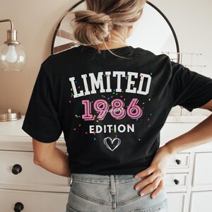 May include: Black t-shirt with the text "LIMITED 1986 EDITION" in white and pink. The design includes confetti and a heart graphic. The shirt is a casual wear item.