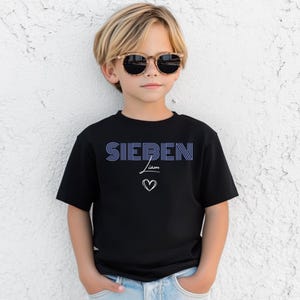 May include: A black short-sleeved t-shirt featuring the word SIEBEN in blue striped lettering, followed by the name Liam in white script and a white outlined heart symbol centered below the text on the front of the garment.