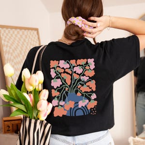 May include: Black t-shirt featuring a floral design on the back. The design includes a blue vase with pink and orange flowers. The shirt is a comfortable, casual style, suitable for everyday wear. The overall theme is floral and botanical.