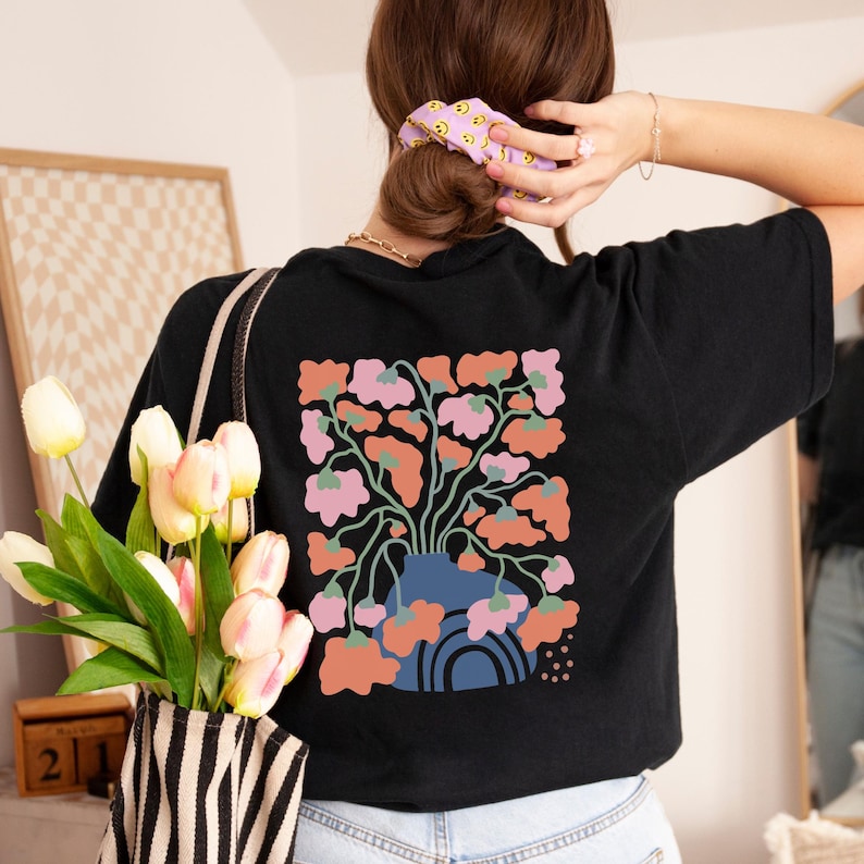 May include: Black t-shirt featuring a floral design on the back. The design includes a blue vase with pink and orange flowers. The shirt is a comfortable, casual style, suitable for everyday wear. The overall theme is floral and botanical.