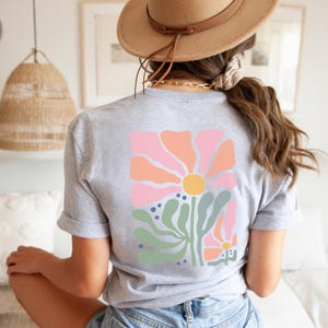 May include: A grey t-shirt with a colourful floral design on the back. The design features a large pink and orange flower with a yellow centre, green leaves, and blue dots. The shirt has rolled-up sleeves.