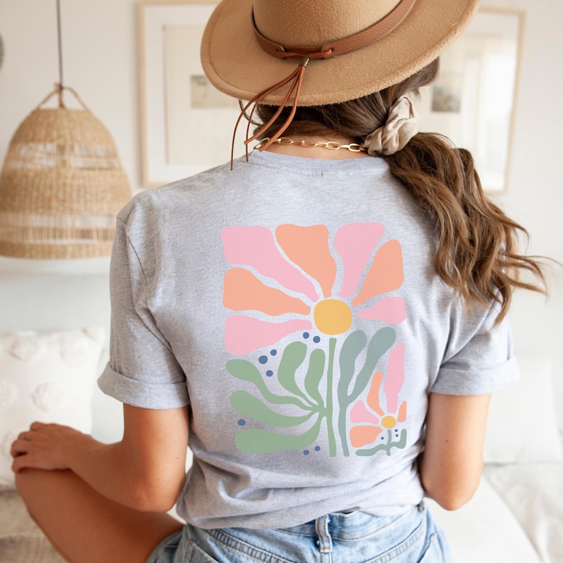 May include: A grey t-shirt with a colourful floral design on the back. The design features a large pink and orange flower with a yellow centre, green leaves, and blue dots. The shirt has rolled-up sleeves.