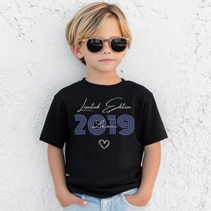Pu&ograve; includere: Una t-shirt nera a maniche corte con la scritta bianca Limited Edition, il numero 2019 a righe blu, il nome Leon in corsivo bianco e il contorno di un cuore bianco. La t-shirt &egrave; indossata con jeans azzurri e occhiali da sole marroni.