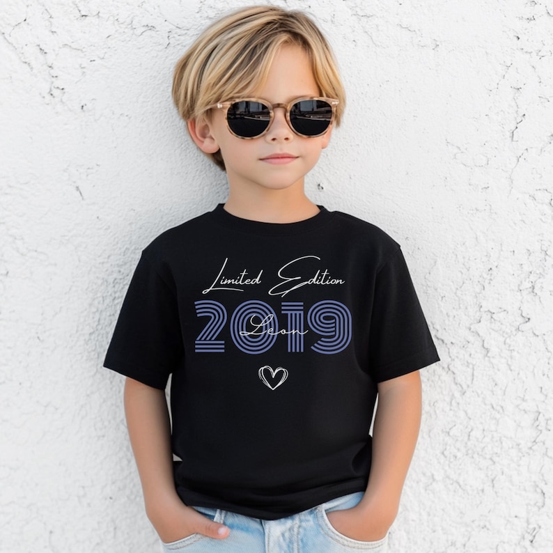 Pu&ograve; includere: Una t-shirt nera a maniche corte con la scritta bianca Limited Edition, il numero 2019 a righe blu, il nome Leon in corsivo bianco e il contorno di un cuore bianco. La t-shirt &egrave; indossata con jeans azzurri e occhiali da sole marroni.