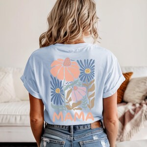 May include: Light blue t-shirt with a floral design in peach, blue, and pink. The word "MAMA" is printed in peach at the bottom of the floral design. The shirt is made of a soft material and has rolled-up sleeves.