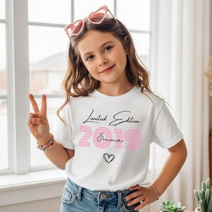 Pu&ograve; includere: Una t-shirt bianca a maniche corte con la scritta Limited Edition in corsivo nero, il numero 2019 in carattere rosa a righe, il nome Emma in corsivo nero e il contorno di un piccolo cuore nero. La maglietta &egrave; abbinata a jeans in denim blu.