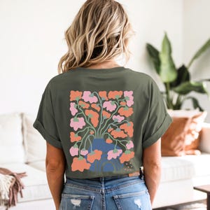 May include: Olive green t-shirt with a floral design on the back. The design features a blue vase with pink and orange flowers. The shirt has rolled-up sleeves and is made of a soft material.