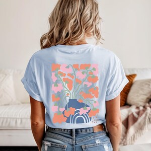May include: Light blue t-shirt featuring a floral design on the back. The graphic includes a blue vase with pink and orange flowers, and a rainbow. The shirt has rolled-up sleeves and a relaxed fit.