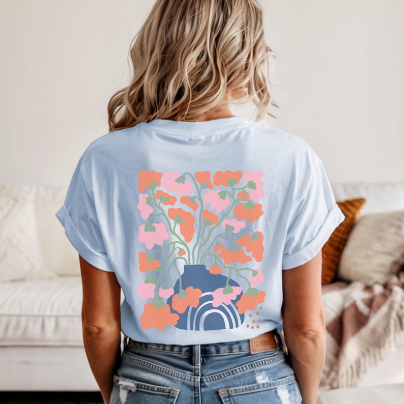May include: Light blue t-shirt featuring a floral design on the back. The graphic includes a blue vase with pink and orange flowers, and a rainbow. The shirt has rolled-up sleeves and a relaxed fit.